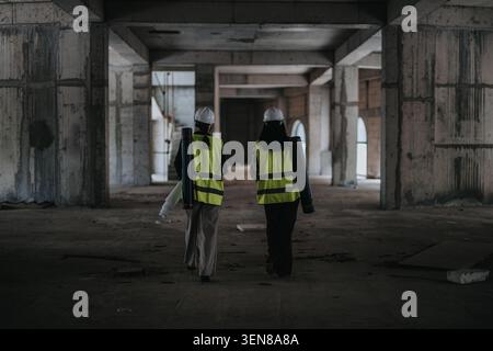 Two female construction professionals walking through unfinished building carrying blueprints and wearing safety gear Stock Photo