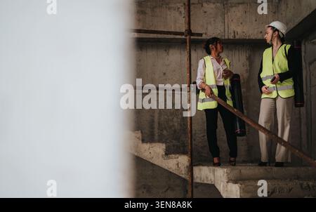 Two female engineers discuss plans on a concrete construction site wearing safety vests and a hard hat Stock Photo