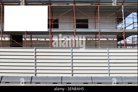 White plastic panel fence in front of building under construction among scffolding. Blank information board on the upper left for copy space. Stock Photo