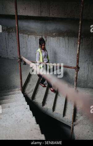 Woman in safety vest carrying blueprint tube down concrete stairs at a construction site in heels Stock Photo
