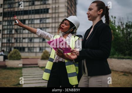 Two female construction engineers discussing site plans wearing hard hat and safety vest near building Stock Photo