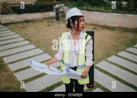 Female architect and engineer on construction site holding blueprints and wearing hard hat and vest Stock Photo