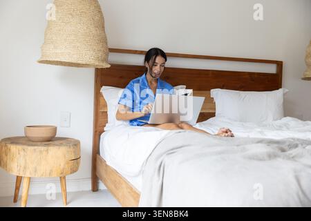 Woman wearing silk pajamas working on silver laptop on wooden-framed bed in minimalist bedroom Stock Photo