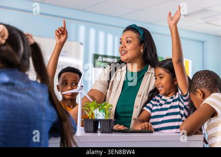 Diverse elementary students with female teacher doing STEM in class with wind turbine plants tablet. Education, curiosity, learning, instructor, scien Stock Photo