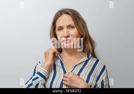 Portrait of smiling mature woman with long hair wearing striped shirt against gray background. Emotional wellbeing, authenticity, optimism. Stock Photo