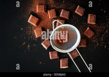 Metal sieve with cocoa powder over chocolate pieces on black background, moody top view baking scene with rich dessert atmosphere Stock Photo