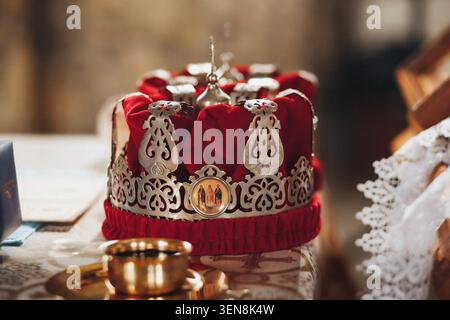 Red velvet crown with ornate silver detailing gleams softly. Golden emblem adorns center, hinting royal legacy. Crown rests on table beside lace and g Stock Photo