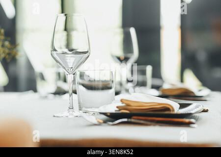 Elegant glassware rests on crisp white linen. Plates, napkins, and cutlery align with precision. Soft light filters through distant windows. Scene sug Stock Photo