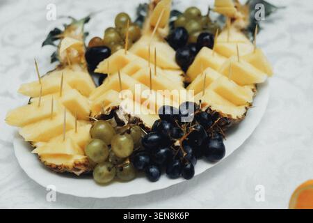 Fresh pineapple slices, skewered with toothpicks, arranged on a plate. Grapes-dark and green-nestle beside the fruit. Plated for serving, it's vibrant Stock Photo