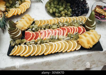 Fruit slices arranged neatly on a dark tray. Bright oranges, grapefruits, and kiwis gleam under soft light. Pineapple pieces and grapes add vibrant co Stock Photo