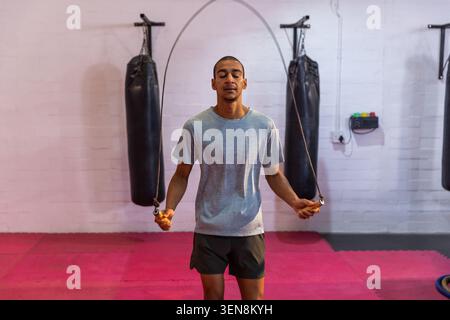 Man jumping rope holding handles on pink mats in gym with heavy bags, wearing gray tee Stock Photo
