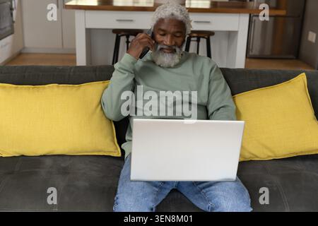 African American senior man talking on phone while using laptop on dark sofa with yellow pillows Stock Photo