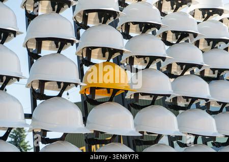 Yellow Hard Hat Standing out from Rows of White Helmets on a Rack. Stock Photo