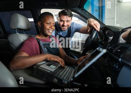 Two mechanics are using diagnostic software on a laptop while sitting in a car at a repair shop Stock Photo