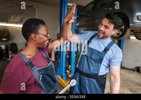 Two smiling mechanics giving high five after successfully repairing a car in a professional auto repair shop Stock Photo