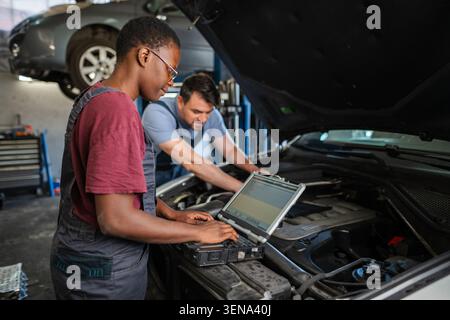 Two auto mechanics are examining a car engine using diagnostic software on a laptop Stock Photo