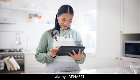 Baking, reading and woman with tablet in kitchen, meal inspiration and search for cuisine tutorial. Home, smile and Asian person with tech for recipe Stock Photo
