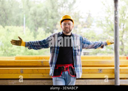 Happy construction worker wearing hard hat and safety goggles, spreading arms in a welcoming gesture on a building site Stock Photo