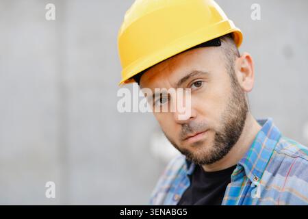 Thoughtful construction worker wearing a yellow hard hat and plaid shirt, concentrating on work at a building site Stock Photo