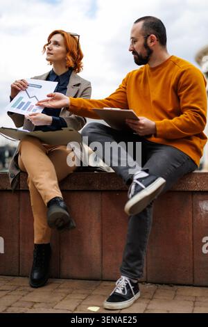 Business meeting outside in a city park with two professionals discussing charts and data on a sunny day while seated on a stone wall Stock Photo