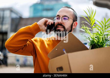 Man talks on phone while holding cardboard box with plants in urban setting during daytime Stock Photo