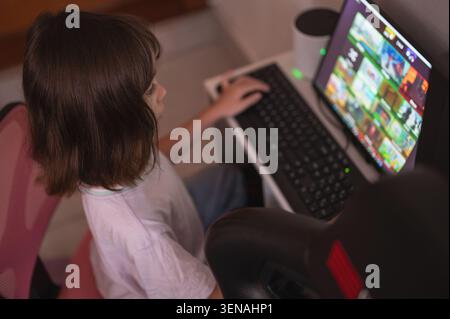 A child sits at a desk using a computer to look for games during the afternoon at home, focused on the screen. Stock Photo