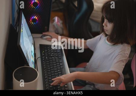 A child sits at a white desk with a computer, focused on playing a game. The room has gaming gear and a relaxed atmosphere. Stock Photo