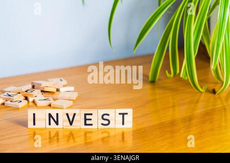 Wooden tiles spelling the word invest next to a spider plant with reflections on the surface and loose tiles behind Stock Photo