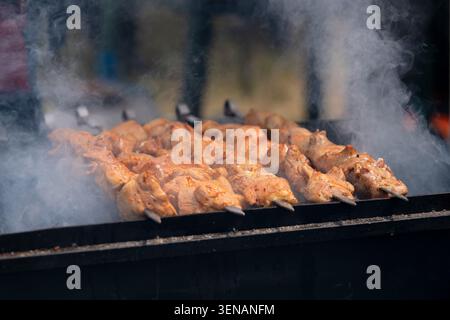 Close-up of seasoned meat on metal skewers grilling over hot charcoal on a black mangal grill, with thick white smoke rising during an outdoor festiva Stock Photo