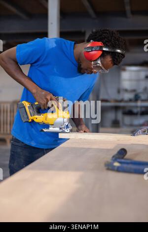 Carpenter is leaning on workshop bench using yellow jigsaw cutting wooden plank under bright lights Stock Photo