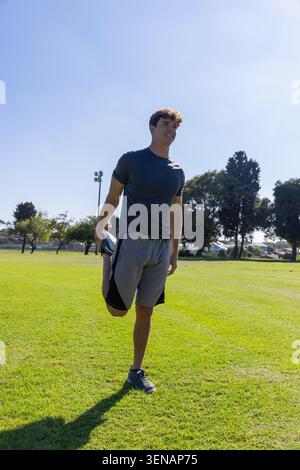 Adult male stretching quadriceps, holding foot on sports field by light pole, wearing running shoes Stock Photo