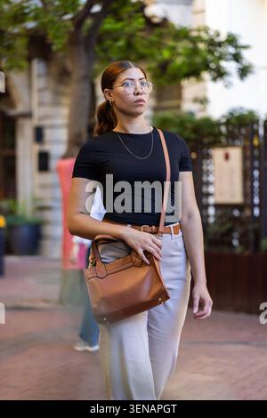 African American woman walking on city sidewalk past shops, wearing black top, carrying brown bag Stock Photo