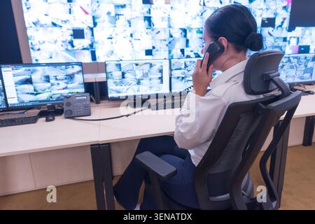 Side view of a female security officer talking on a landline telephone while monitoring multiple live camera feeds on screens in a high-tech control c Stock Photo