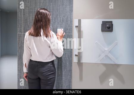 Rear view of a female security guard in uniform operating a large steel vault door wheel, using high-security access keypads to open a secure deposito Stock Photo