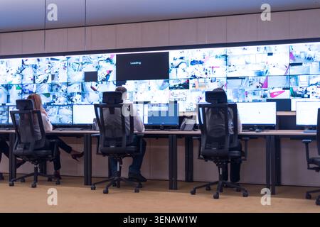 Wide-angle view of multiple security operators sitting at desks in front of a massive video wall displaying dozens of live closed-circuit television ( Stock Photo