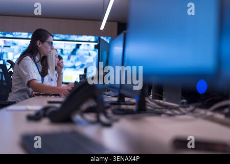 Side profile view of professional female operators sitting in a row, using computers to monitor security feeds with large multi screen video walls vis Stock Photo