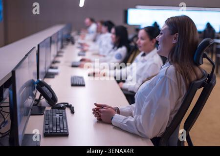 Side perspective of a long row of female dispatch operators wearing white uniforms, sitting at computer desks and smiling while working in a bright se Stock Photo