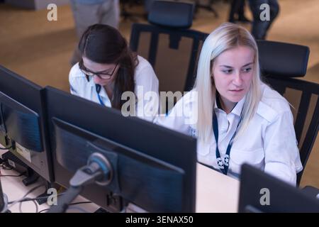 Two professional female security operators in white uniform shirts sitting at desks and focused on computer monitors in a high tech surveillance contr Stock Photo
