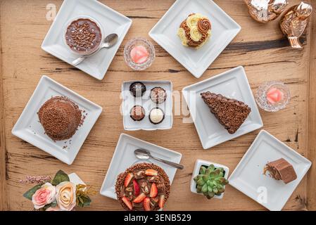 Overhead shot featuring brigadeiros, cake slice, pudding, and succulent arranged on small white plates over a light brown wooden table surface. Select Stock Photo
