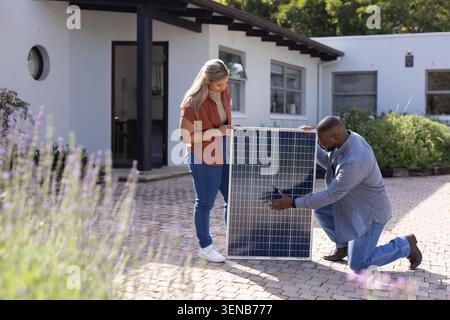 African American woman holding solar panel in rust shirt while man tech kneeling on brick driveway Stock Photo