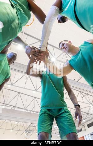 Diverse male teammates huddling on basketball court, stacking hands over camera in green jerseys. Lowangle, athletic, teamwork, huddle, unity, focus, Stock Photo