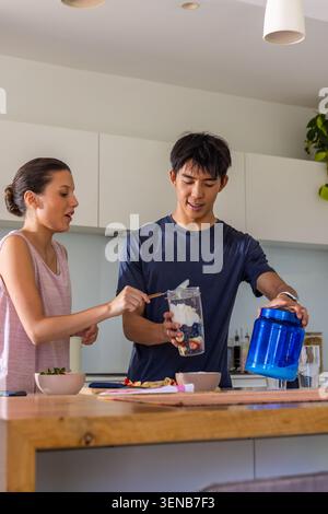 Diverse couple preparing berry yogurt smoothie at wood island, using blender cup and blue can Stock Photo