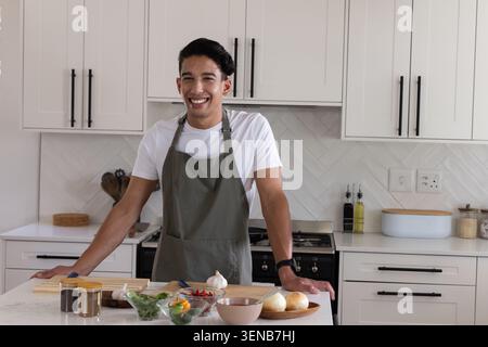 African American man wearing gray apron, leaning on kitchen island, preparing salad with spice jars Stock Photo