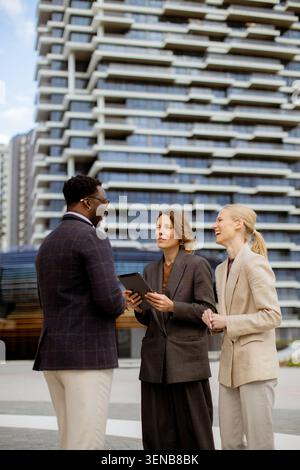 Three individuals stand together in an urban area. They are engaged in a discussion while checking a tablet. The background shows modern buildings and Stock Photo