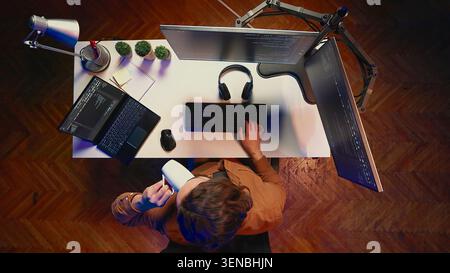 Top-down view of engineer doing multitasking to finish developing PC code, drinking coffee, listening music. IT admin wearing headphones, solving database errors on computer and laptop, aerial shot. Stock Photo