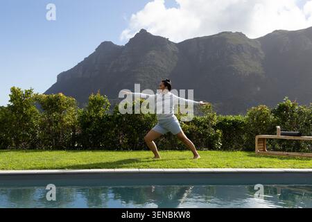 Mid-30s Asian woman practicing yoga on lawn beside pool, wearing gray hoodie with water bottle near Stock Photo