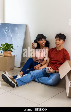 Diverse couple sitting on tiled floor in new home taking break from unpacking boxes holding mugs Stock Photo