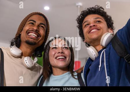 Diverse friends posing and smiling while taking selfie in lobby with headphones and beanie Stock Photo