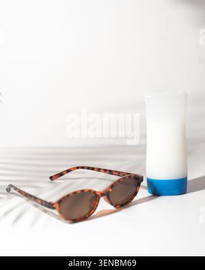 Sunglasses and sunscreen bottle on a white background with tropical palm leaf shadows. Clean summer still life for beach, sun protection, skincare and Stock Photo