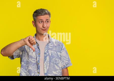 Middle-aged man showing thumbs down with frown and strong disapproving look, expressing rejection, disagreement and dissatisfaction. Young guy giving negative gesture isolated on yellow background. Stock Photo